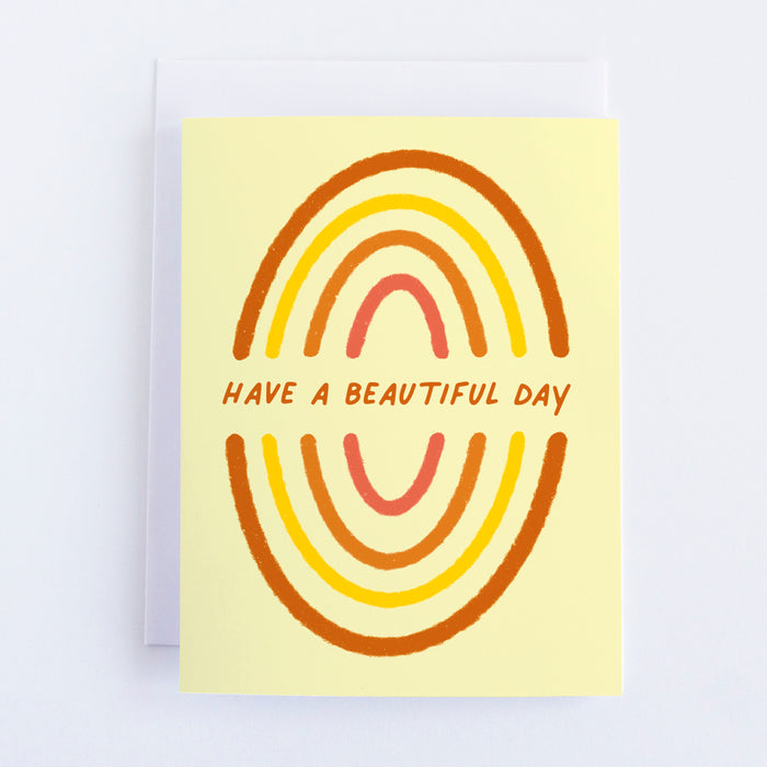 A yellow card with two mirrored rainbows painted in orange and yellow and pink and in the middle of the rainbows the text reads Have a Beautiful Day.