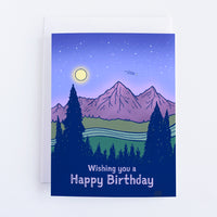 A blue card with a night sky and a purple mountain landscape with a shooting star above the mountains. The Forest in the foreground is dark blue and the text in the forest area says Wishing you a Happy Birthday.