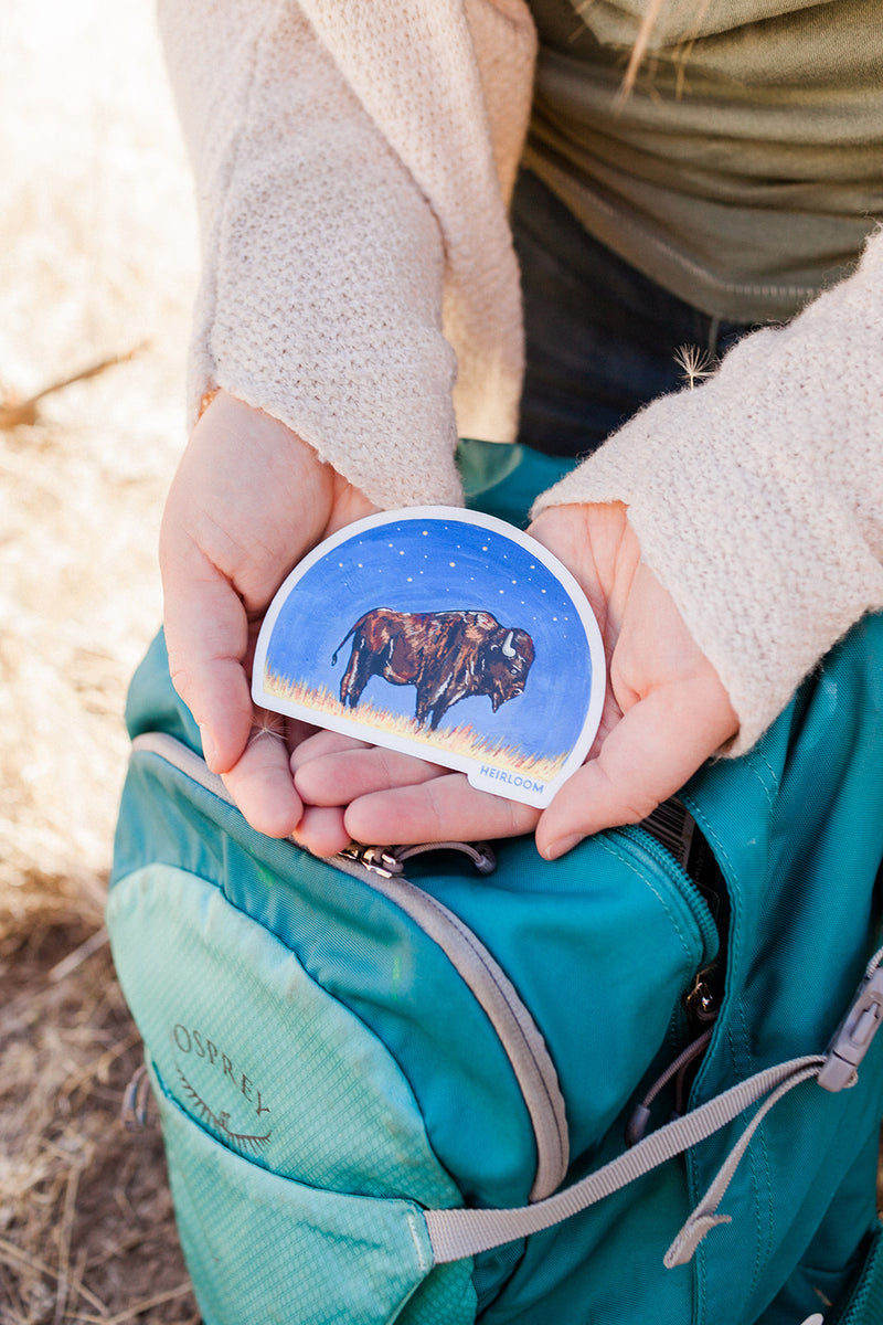 Woman holding a vinyl sticker featuring a bison in profile against the backdrop of a night sky. It is a hand painted design that has been reprinted on a durable vinyl sticker. it is a half circle sticker. 
