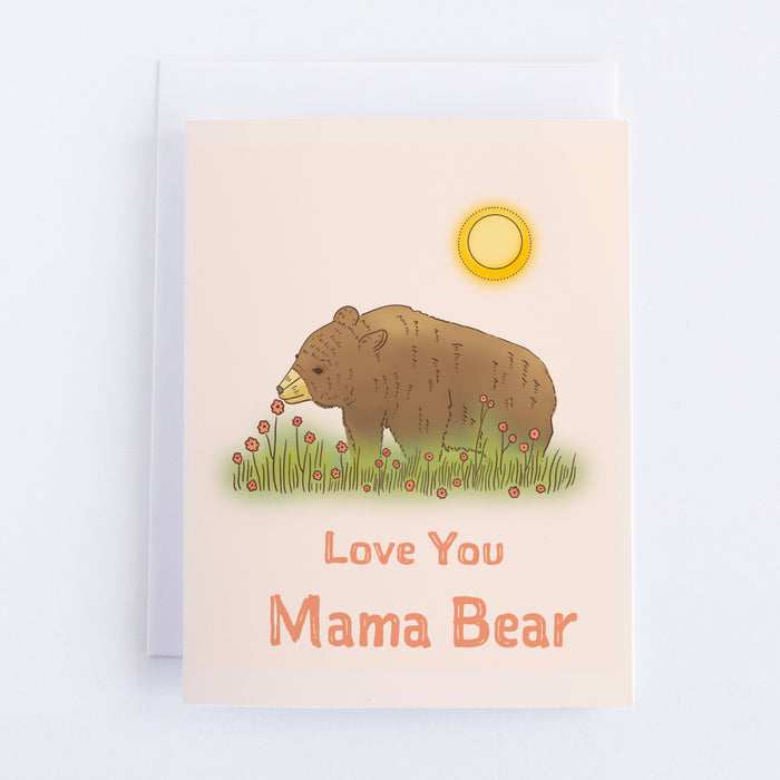 A light pink card with a small brown bear in a field sniffing a pink flower. It is surrounded by small pink flowers. The text below reads Love You Mama Bear.