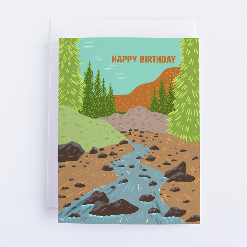 A greeting card with a mountain river and mountains and trees in the background and the text at the top of the card says Happy Birthday.