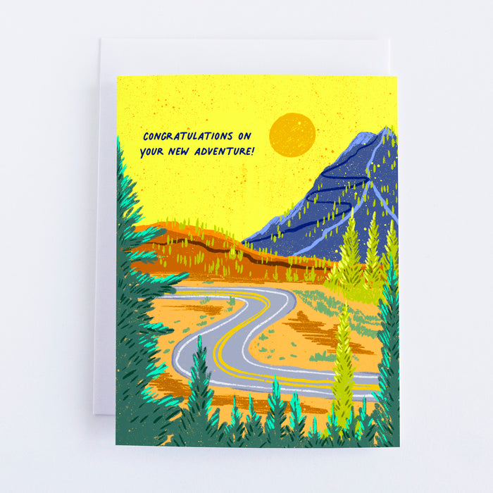 A yellow greeting card with a mountain road and mountains in the background and trees in the foreground. The text says Congratulations on your new adventure.