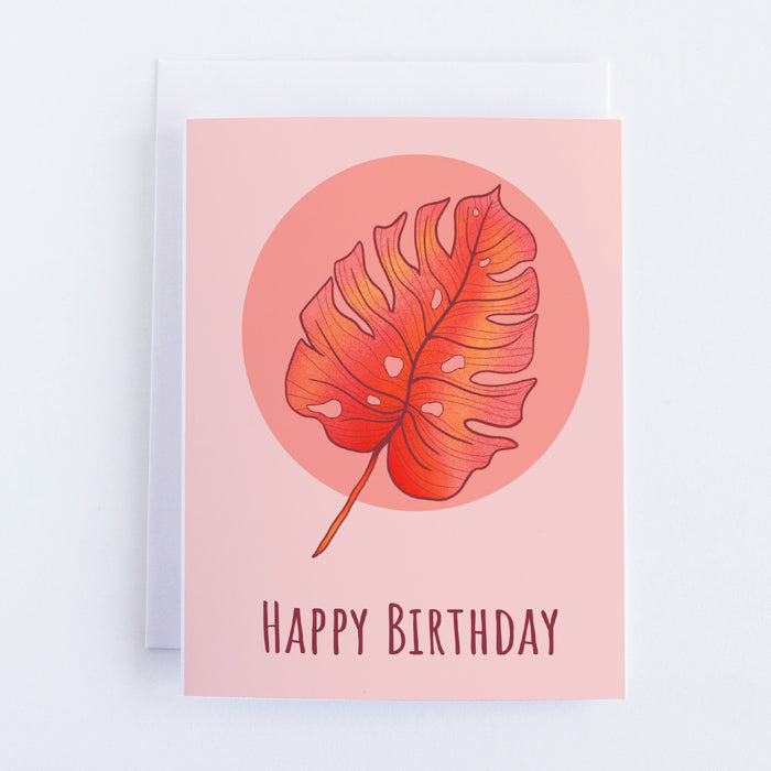  A pink greeting card with a dark pink circle on which sits a pink, orange, and yellow monstera leaf. The text underneath it reads happy birthday.