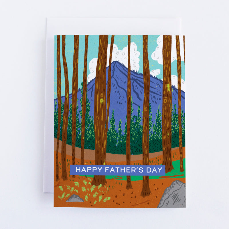 A greeting card with a mountain in the background and tree trunks in the front and a banner in the foreground that says Happy Fathers Day.